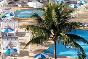 Un espace piscine doté de plusieurs chaises longues et de parasols, d’un palmier et d’une piscine à l’eau d’un bleu limpide.