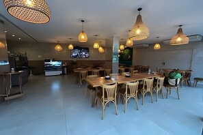 Un vaste espace dédié à la salle à manger, avec de nombreuses tables et chaises, des luminaires suspendus et un grand écran de télévision en arrière-plan.