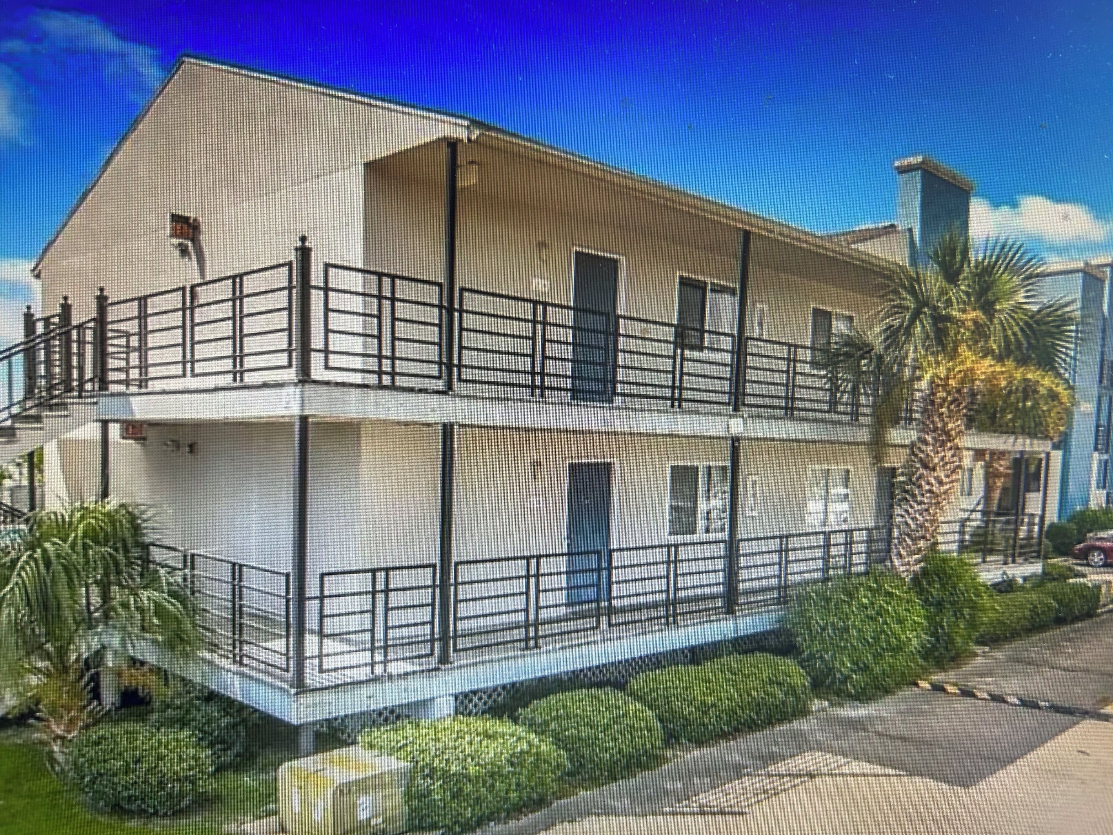 A two-story building with balconies, a flat roof, and a small garden area.