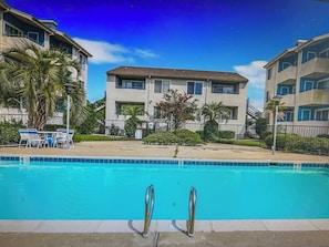 Una piscina con agua cristalina, rodeada por una valla, junto a un edificio de varios pisos con balcones.