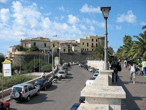 A street with parked cars, a pedestrian walkway, and buildings in the background.
