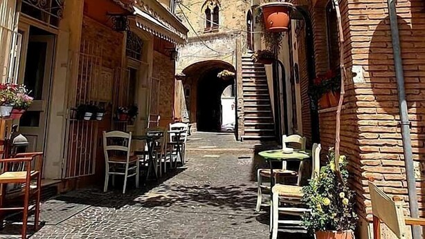 A narrow street with buildings on either side, outdoor seating, and a brick tower in the background. - caty house (Nettuno)