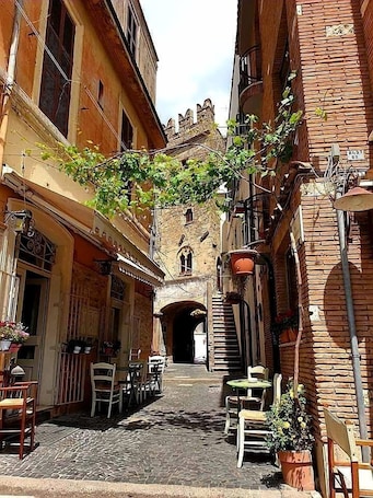 A narrow street with buildings on either side, outdoor seating, and a brick tower in the background.. caty house