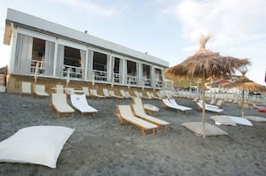 A beachside hotel with rows of sun loungers, a thatched umbrella, and a building with large windows.