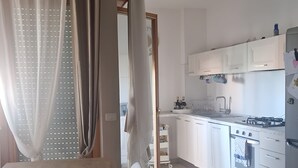 A modern kitchen with white cabinets, a dining table, and a view into the adjacent room. - caty house (Nettuno)
