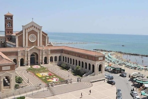A church with a clock tower, a courtyard with a flower bed, and a beach in the background.