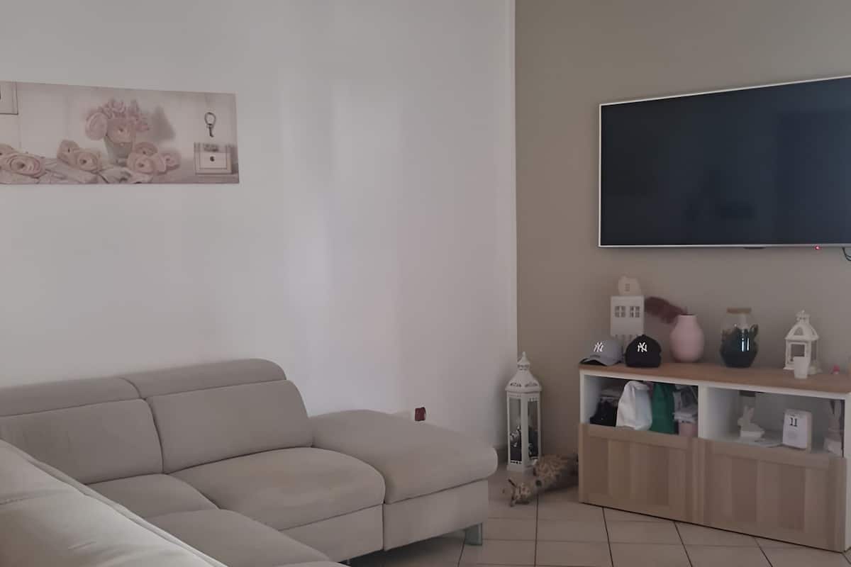 A living room with a beige sectional sofa, a flat-screen TV mounted on the wall, a small side table, and a shelf with various decorative items.