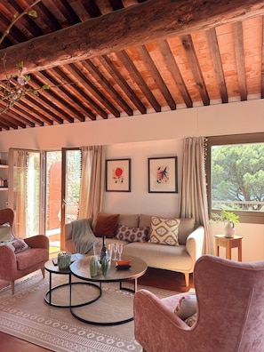 Ein gemütliches Wohnzimmer mit Holzbalkendecke, einer Sofagarnitur, einem Couchtisch und Blick durch große Fenster.