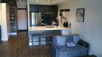 A modern kitchen with a central island, dark cabinetry, stainless steel appliances, and a grey sofa in the living area.