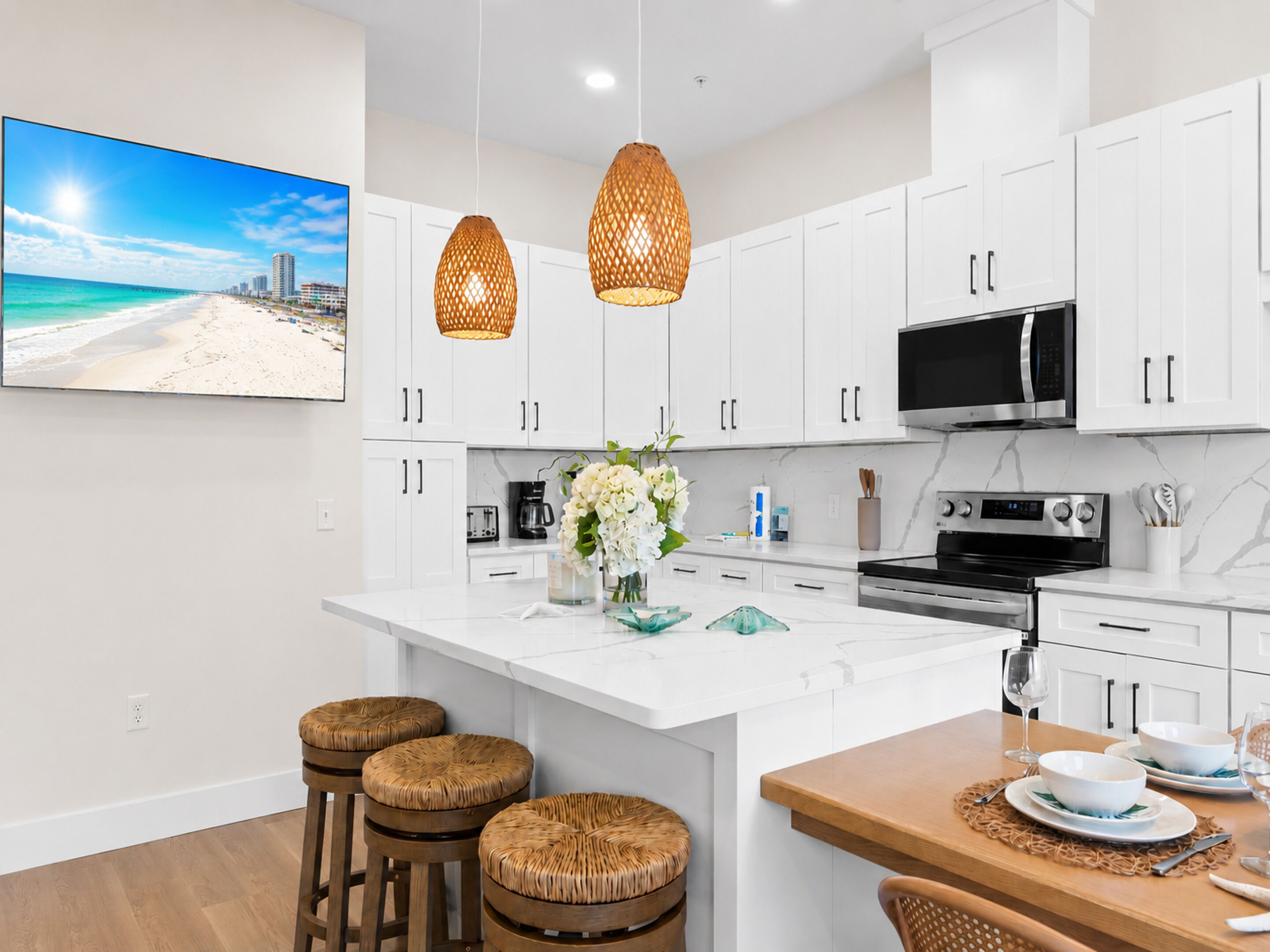 A modern kitchen with white cabinets, a large island, wicker stools, a TV, and a beach view.