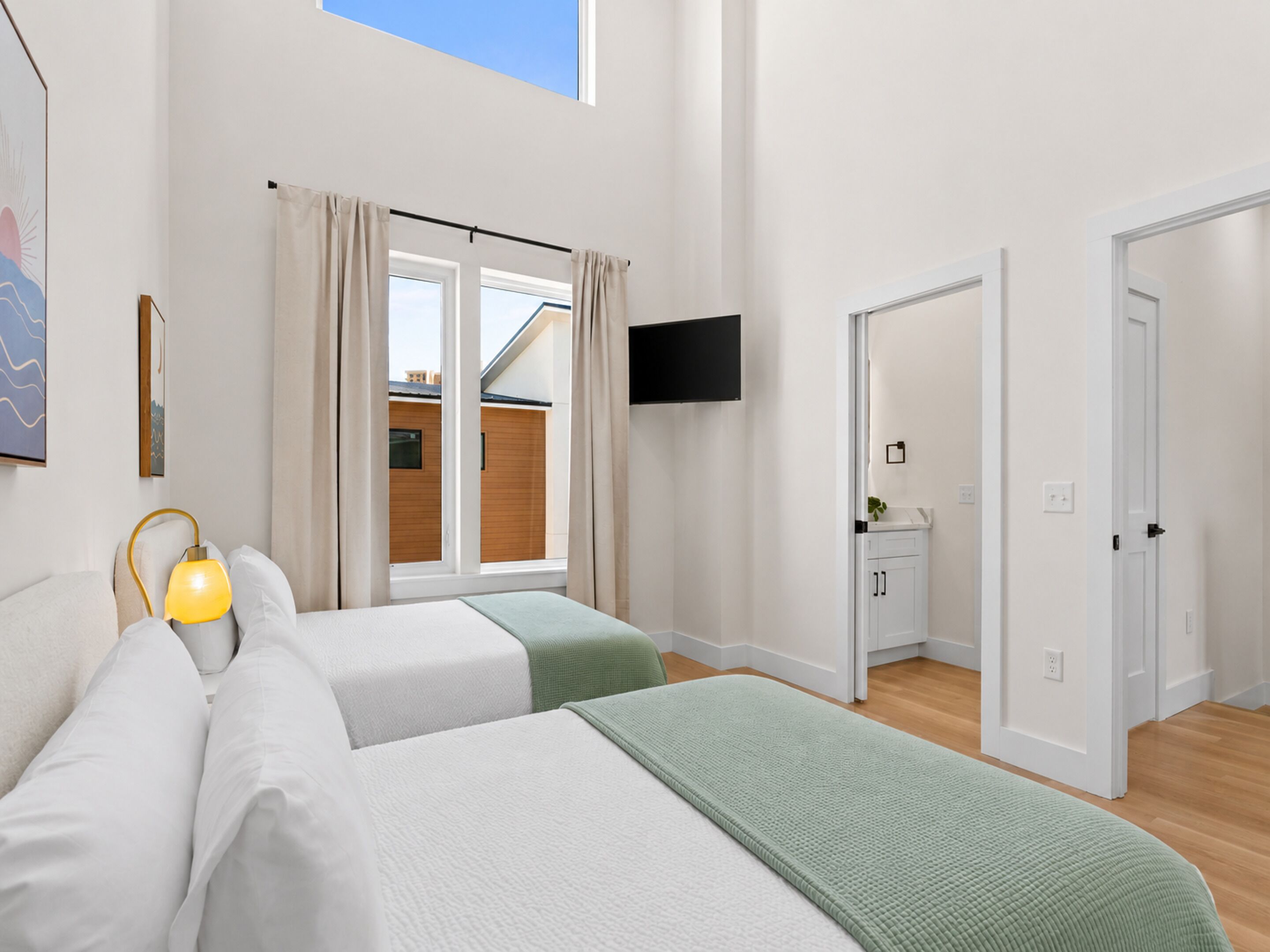 A bedroom with two beds, a TV, a window with curtains, and a door leading to a bathroom.