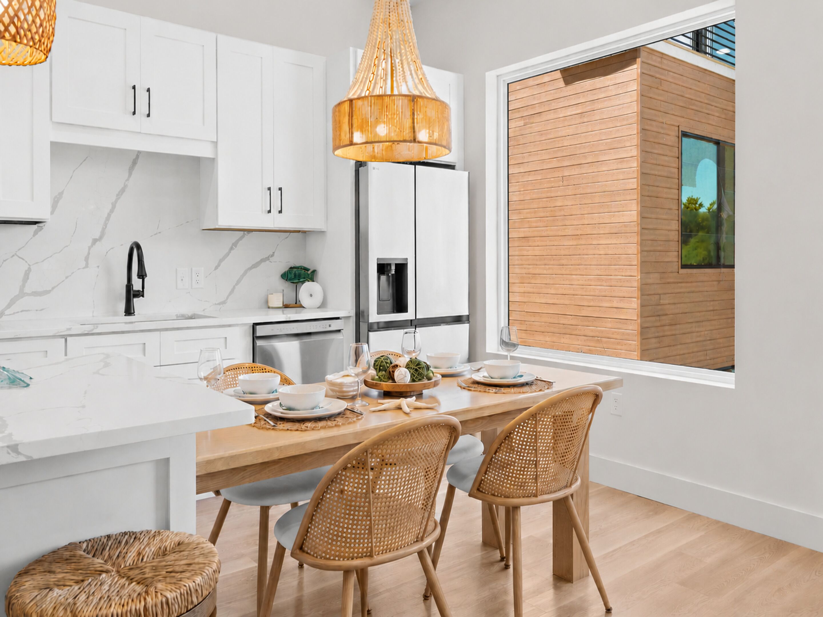 A modern kitchen with white cabinets, a wooden dining table, wicker chairs, and a large window.