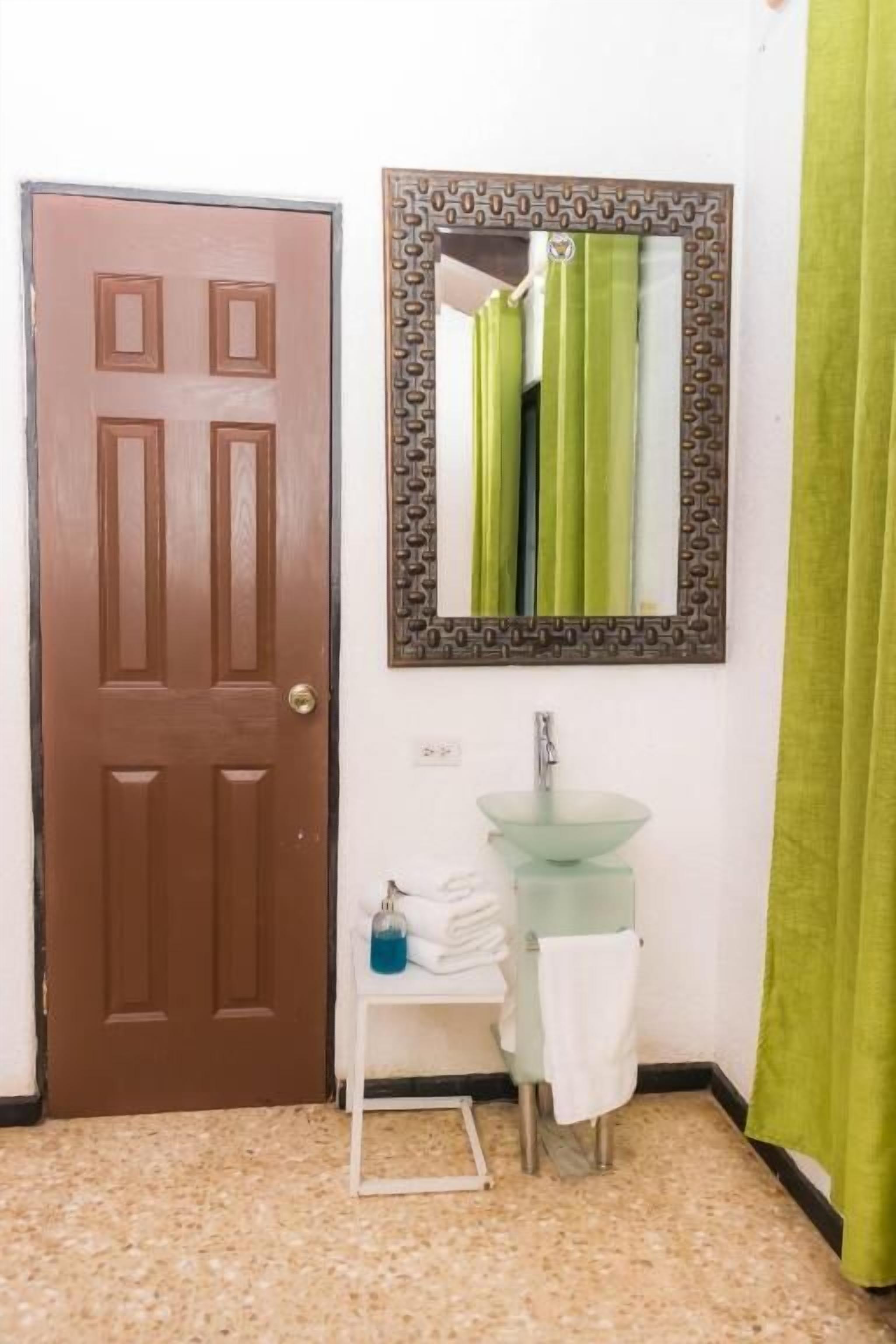 A bathroom with a brown door, a glass sink, a mirror with a patterned frame, and a green curtain.