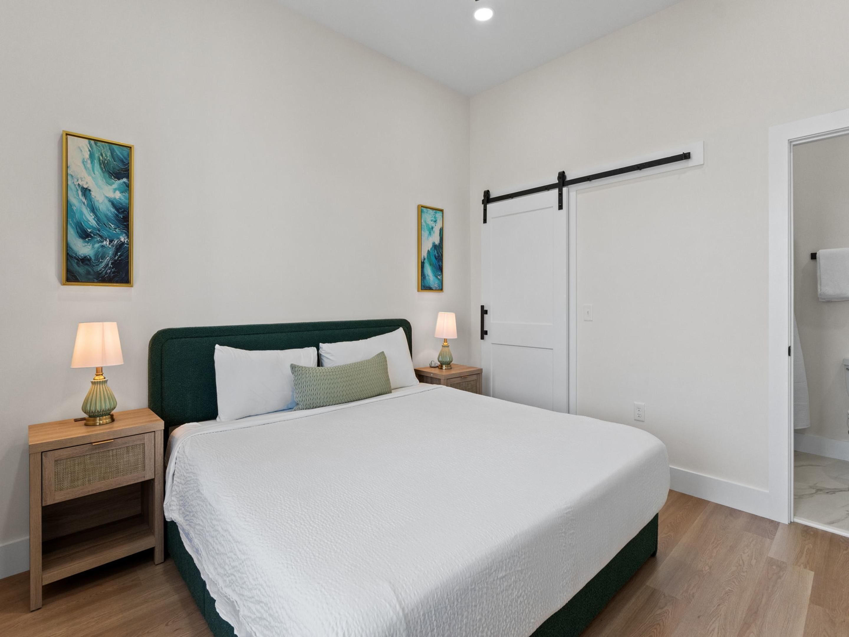 A bedroom with a bed, two bedside tables, a nightstand, a door, and two wall-mounted paintings.