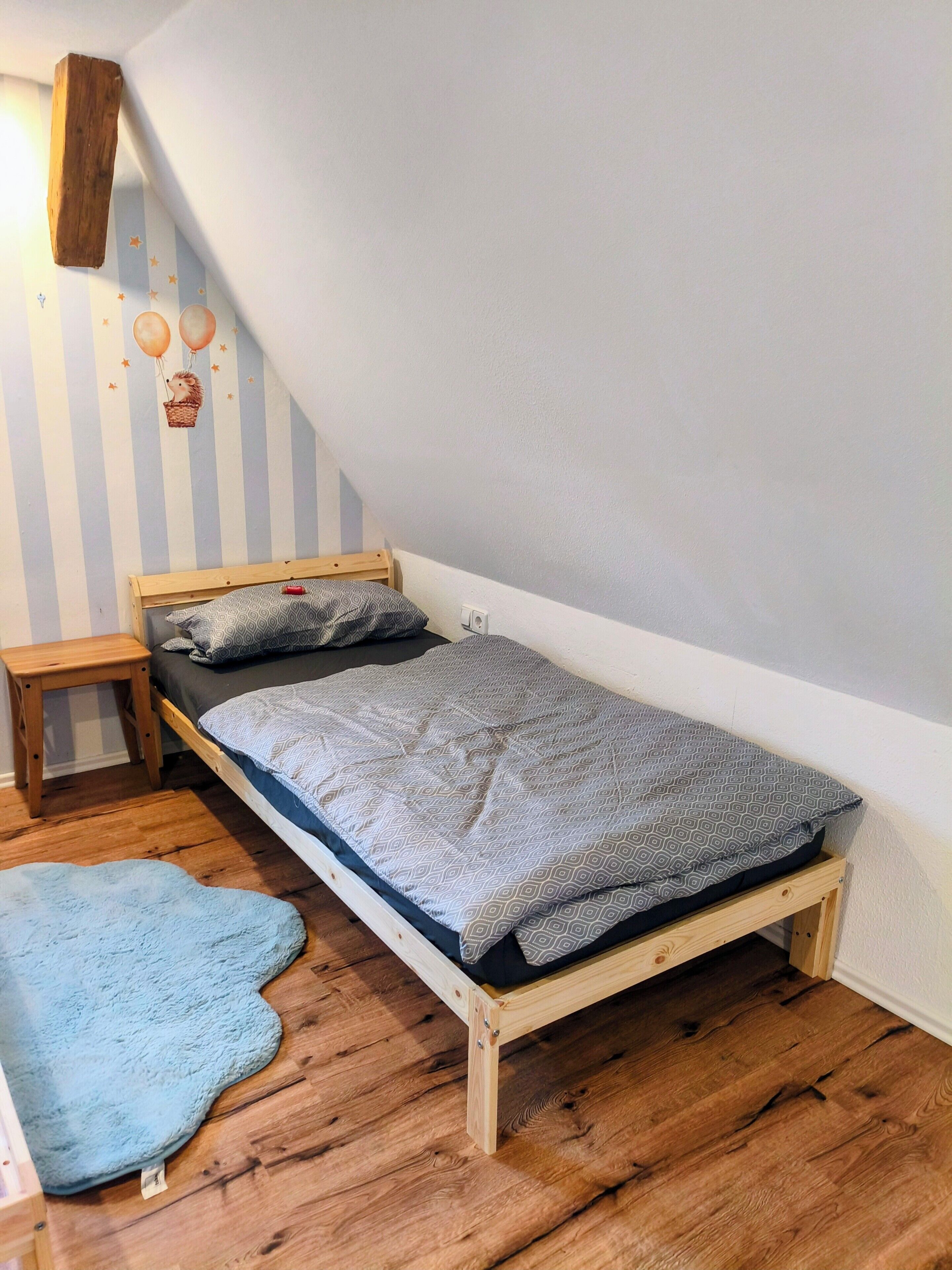 A small, single-bed room with a sloped ceiling, a wooden bed frame, a bedside table, a blue rug, and a wall with a striped pattern and a wall-mounted shelf.