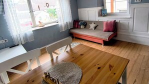 A room with a wooden floor, a white sofa with red cushions, a white chair, a woven rug, and a window with sheer curtains. - Vacation cottage Obermarchtal (Obermarchtal)