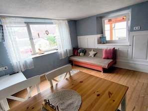A room with a wooden floor, a white sofa with red cushions, a white chair, a woven rug, and a window with sheer curtains.