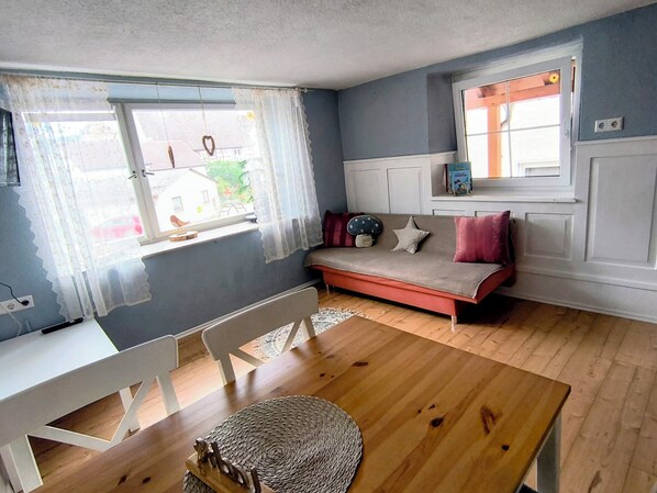 Une pièce avec un plancher en bois, un canapé blanc orné de coussins rouges, une chaise blanche, un tapis tissé et une fenêtre avec des rideaux légers.