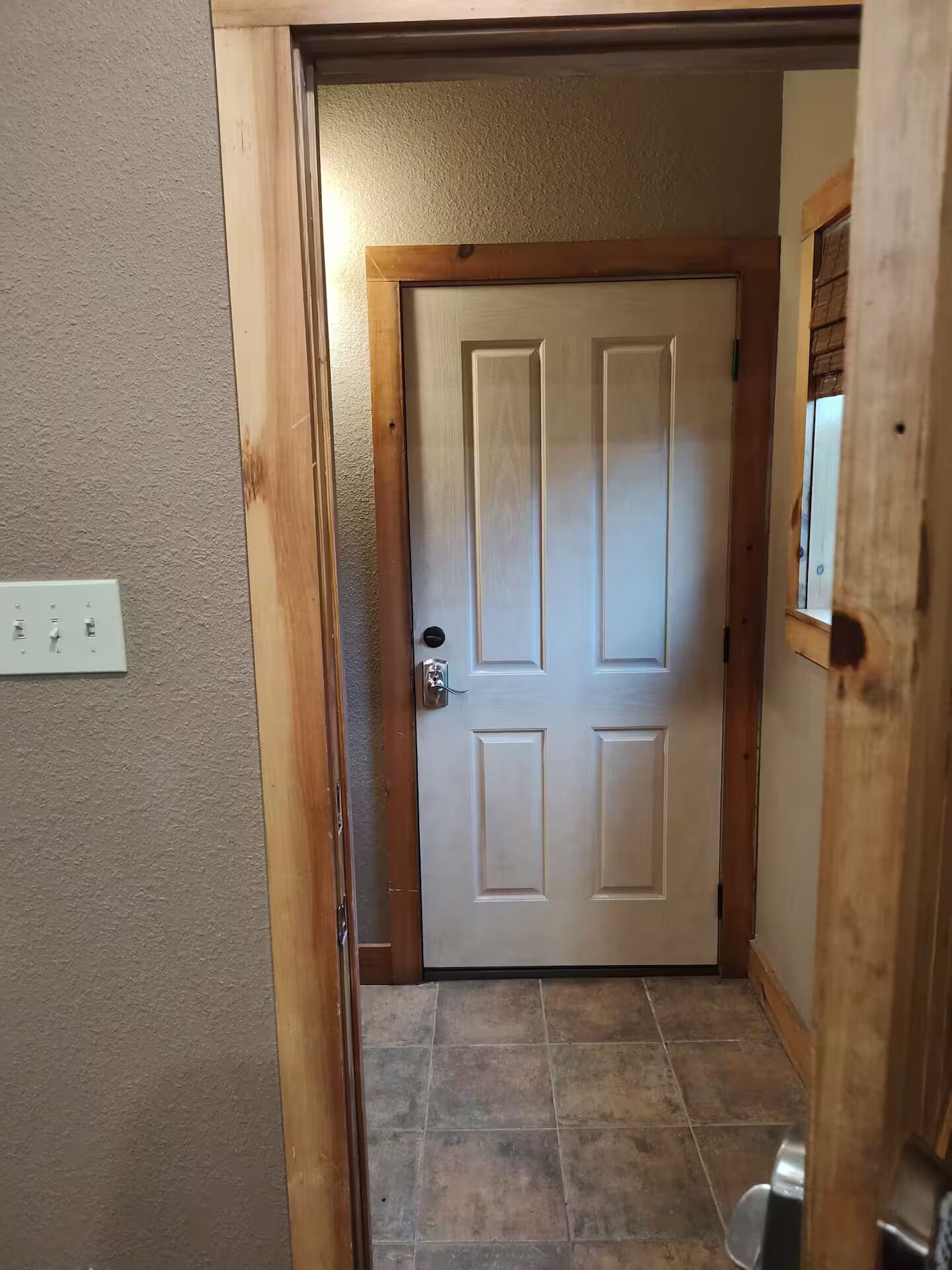 Un pasillo con puerta de madera, un interruptor de luz en la pared y suelo de baldosas.