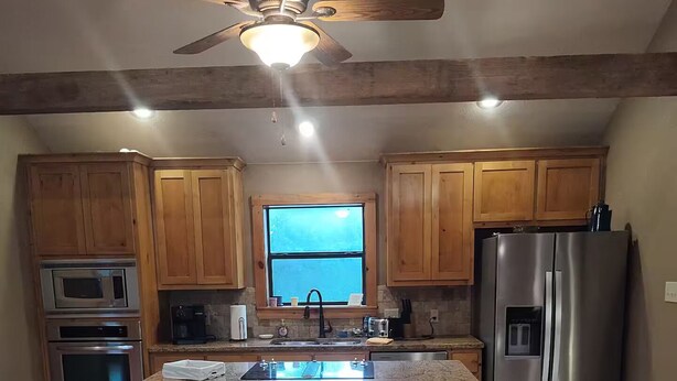 A kitchen with wooden cabinets, a stainless steel refrigerator, a microwave, an oven, a ceiling fan, and a window. - The Farm House at Bliss Point Ranch FM-17 (Grand Saline)