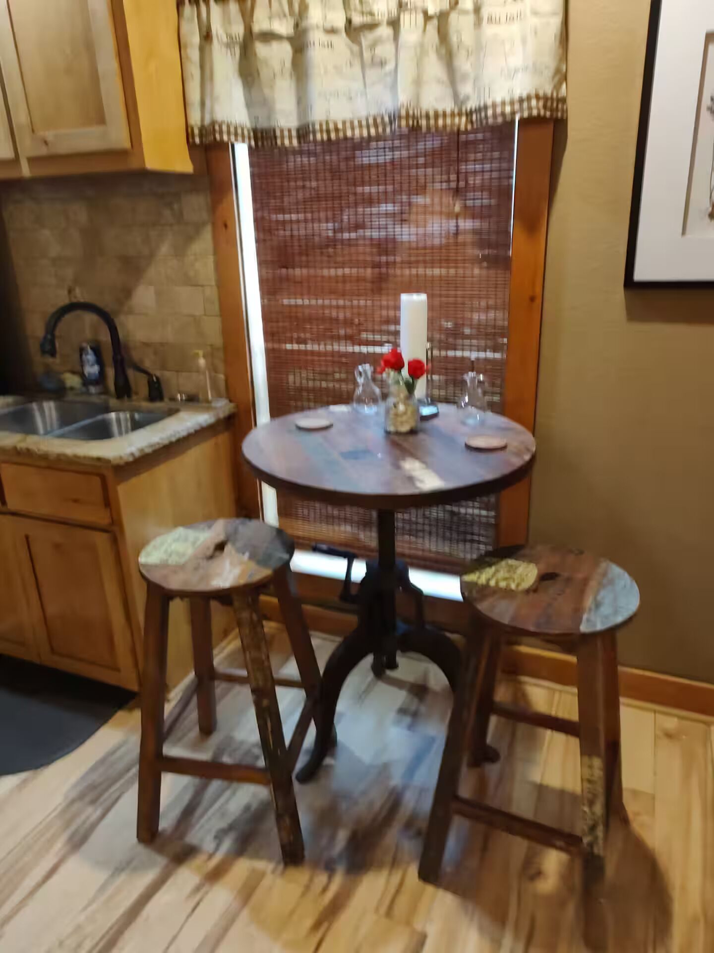 A kitchen with wooden cabinets, a round table with stools, a window with a patterned curtain, and a brick wall.