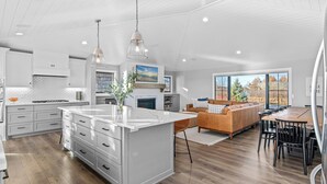 Una cocina moderna con armarios blancos, una isla central, iluminación colgante y un comedor con mesa y sillas de madera.