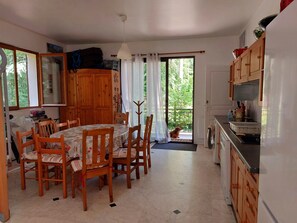 A kitchen with wooden cabinets, a dining table with chairs, a fan, and a view through the door. - The 3 Pignons gourmet refuge (Noisy-sur-École)