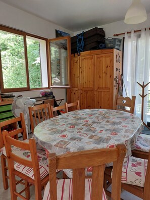 A dining area with a round table covered by a patterned tablecloth, wooden chairs, a wooden cabinet with hooks, and a window with a view of greenery. - The 3 Pignons Gourmet Retreat (Noisy-sur-École)