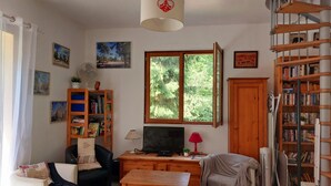 A living room with a sofa, a TV, a wooden desk, a bookshelf, a window with a view of trees, and a spiral staircase. - The 3 Pignons gourmet refuge (Noisy-sur-École)