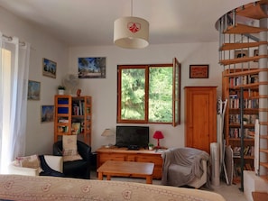 A living room with a sofa, a TV, a wooden desk, a bookshelf, a window with a view of trees, and a spiral staircase. - The 3 Pignons gourmet refuge (Noisy-sur-École)
