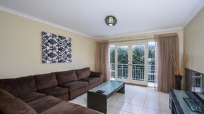 Ein Wohnzimmer mit einem braunen Sofa, einem Glastisch, einem Fernsehmöbel und einem großen Fenster mit Vorhängen.