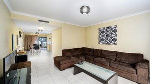 Ein Wohnzimmer mit einem braunen Ecksofa, einem Glastisch und einem Fernseher auf einem Schrank. Es gibt einen Essbereich mit Tisch und Stühlen, sowie ein großes Fenster mit Vorhängen.