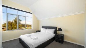 Ein Schlafzimmer mit großem Fenster, einem Bett, einem Nachttisch und einer Lampe.