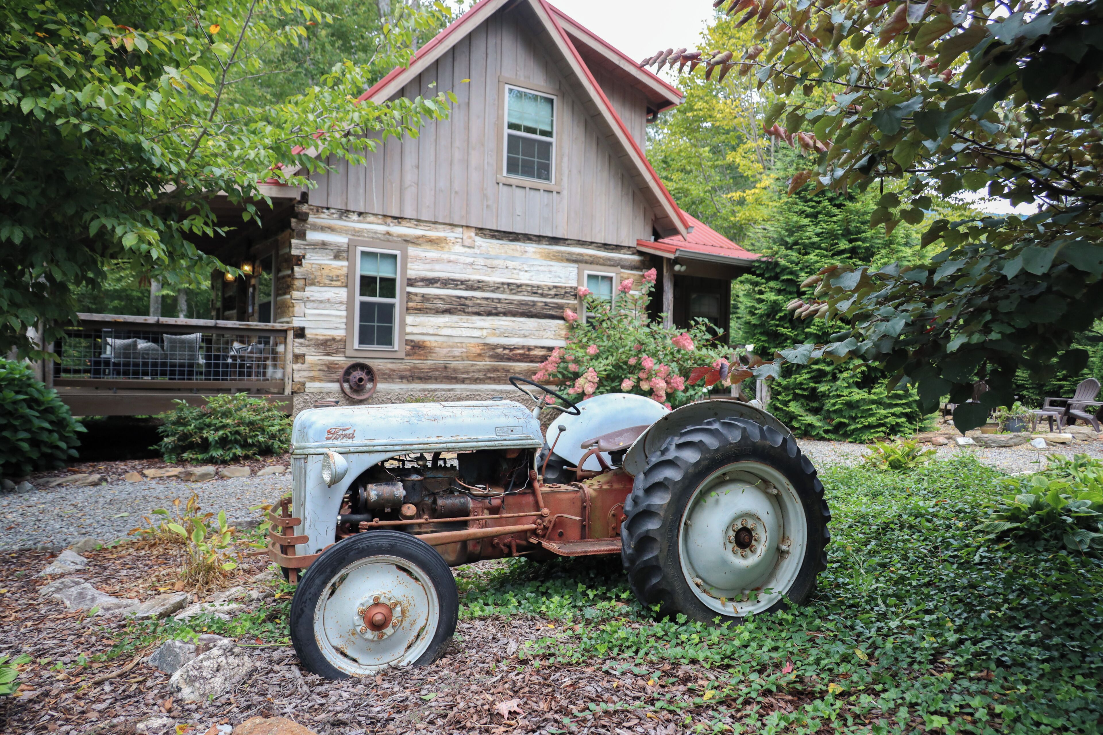A rustic wooden house with a red roof, a vintage tractor parked in front, and a gravel driveway.