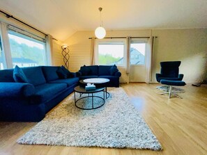 Een woonkamer met een hoekbank, een ronde salontafel, een klein bijzettafeltje en een hoge stoel. Er zijn ramen met gordijnen, een hanglamp en een houten vloer.