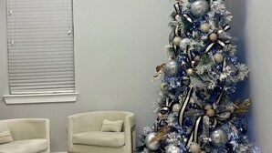 Una stanza con pavimento a scacchi, due poltrone bianche, un albero di Natale decorato con ornamenti argentati e dorati e una finestra con persiane.