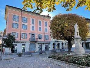 Ein öffentlicher Platz mit einem rosafarbenen Gebäude, Bänken, einer Statue und einem Baum mit Herbstlaub.