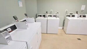 A laundry facility with multiple washing machines and dryers, a tiled floor, and a plain wall with a small sign. - Labor Day Opening🌴Last minute PCB Getaway  The Pink Panama at Origin at Seahaven (Panama City Beach)