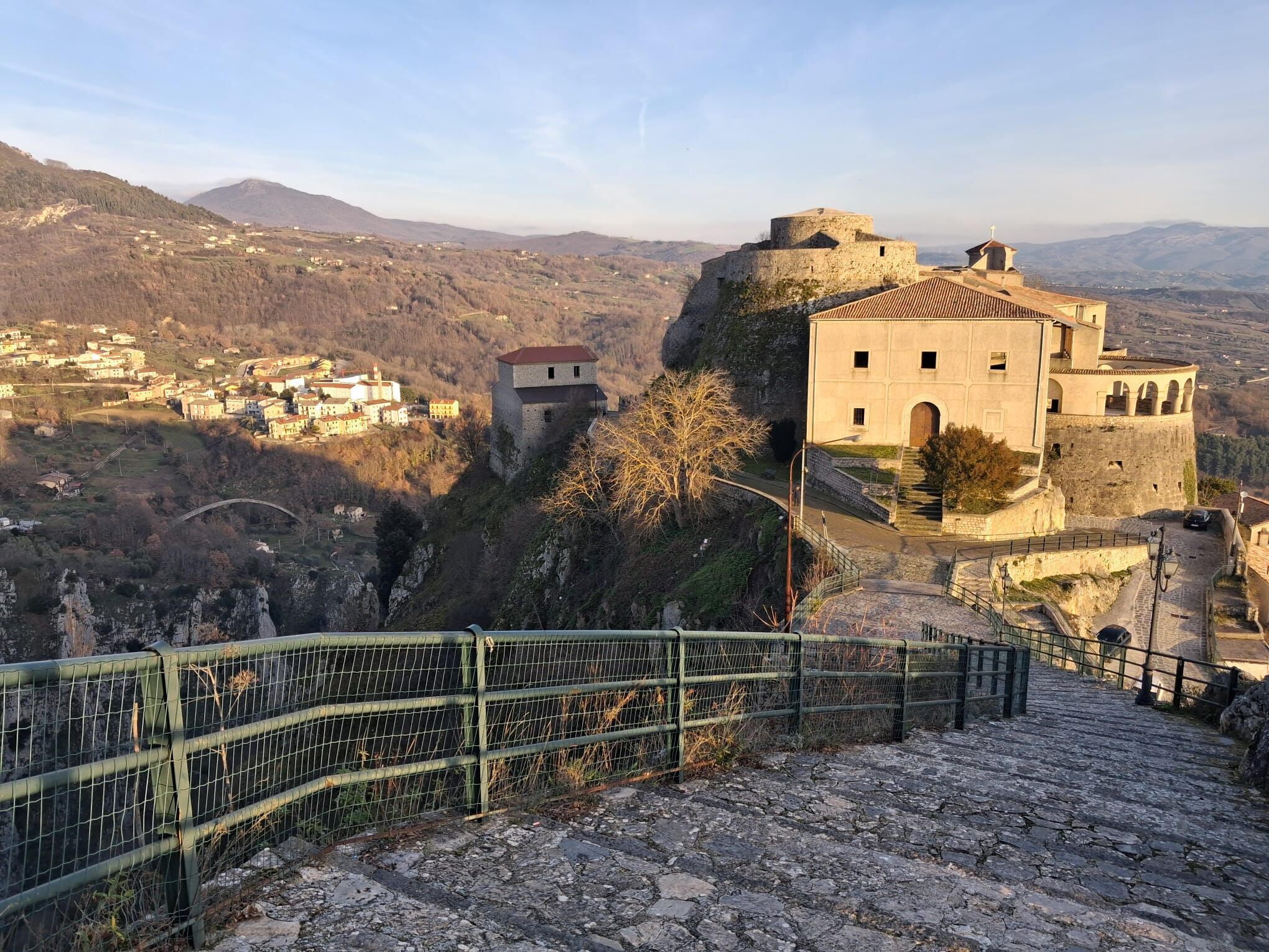 Ein historisches Steinhaus mit einem Turm, auf einem Hügel gelegen und mit Blick über ein Tal mit verstreuten Häusern.
