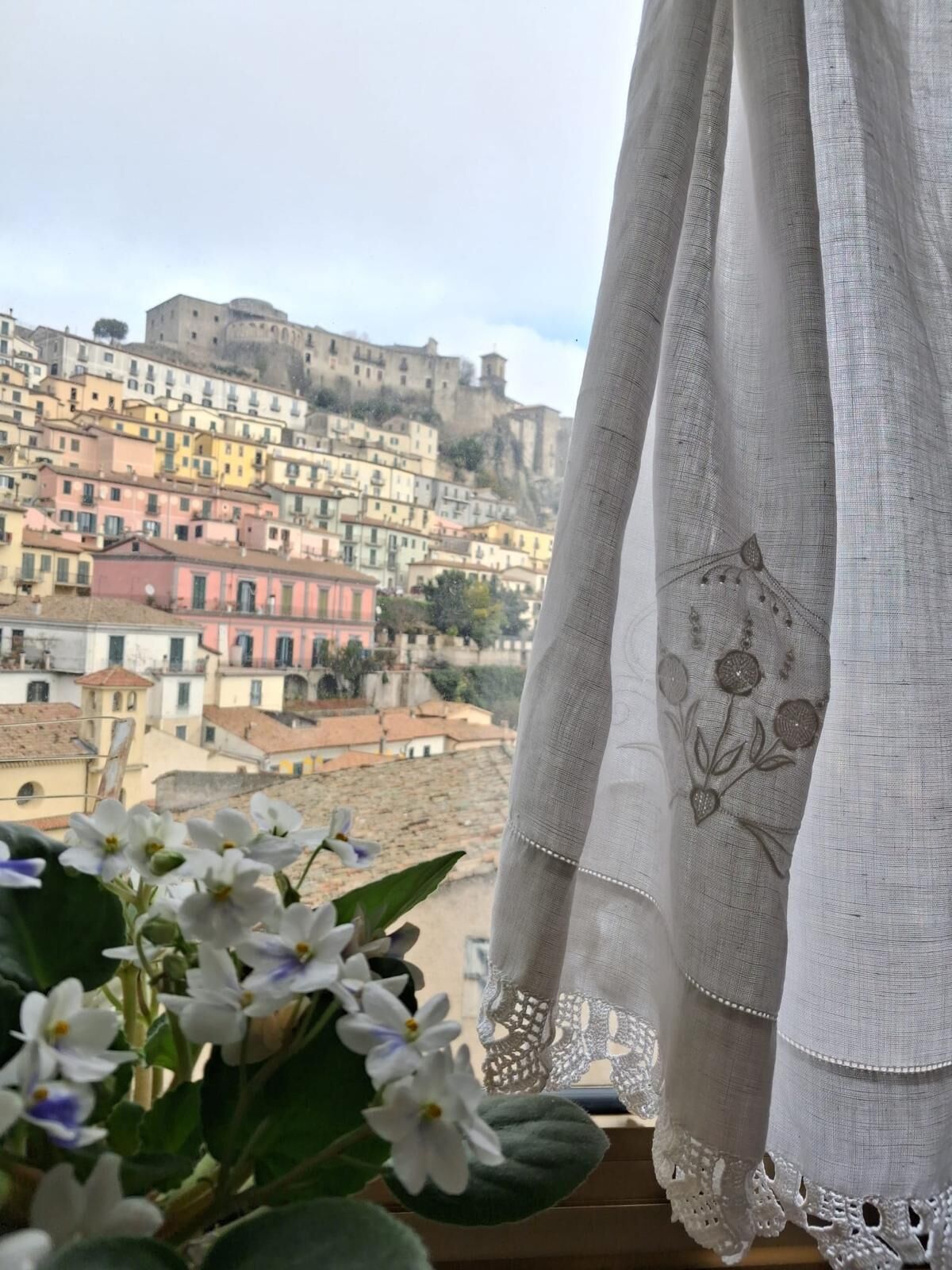 A window with a view of a hillside town, a white curtain with floral embroidery, and a potted plant with white flowers.