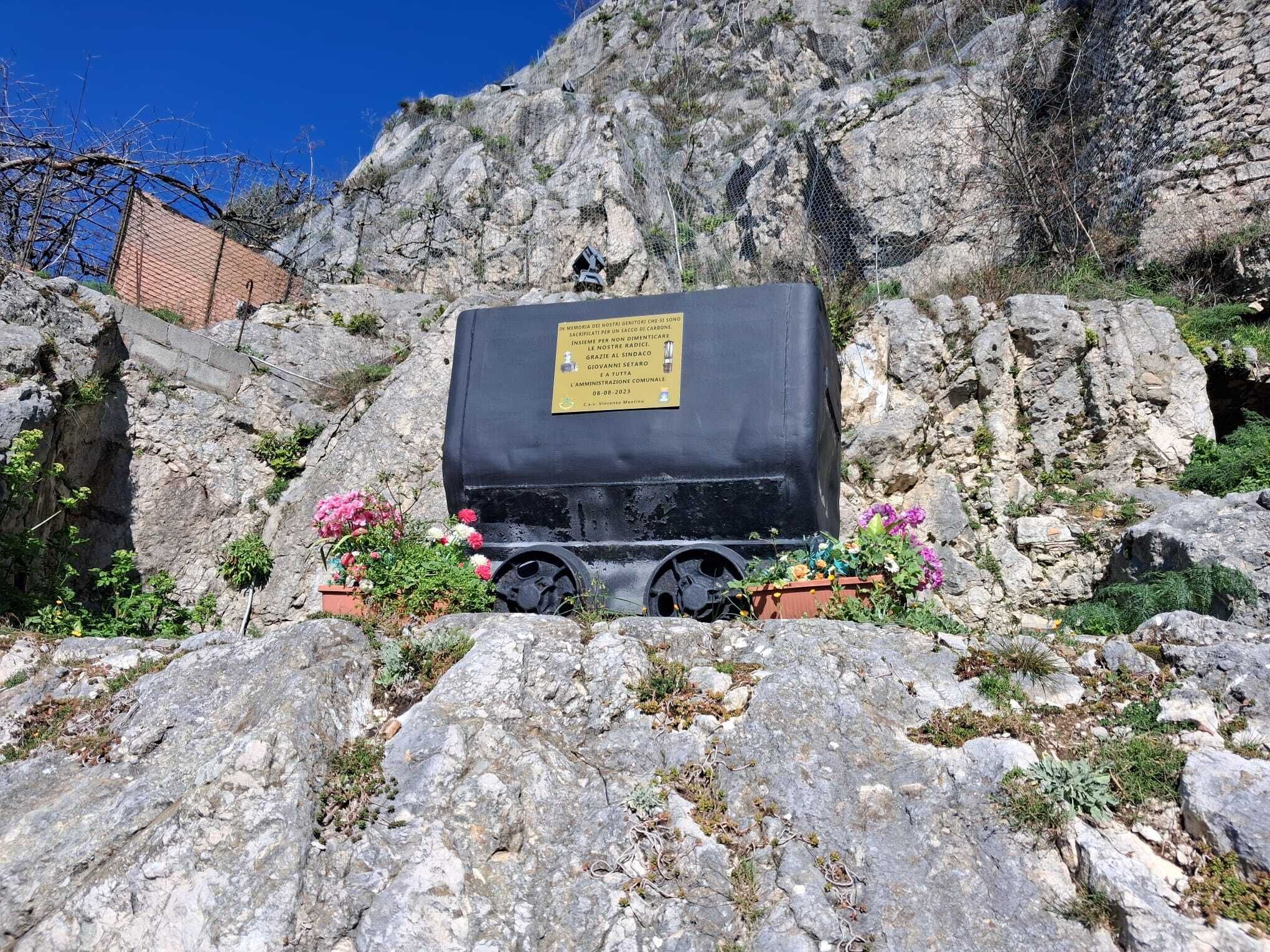 A memorial with a black container and a plaque, surrounded by rocky terrain and flowers.