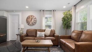 A living room with a brown leather sofa, a wooden coffee table, a round wall clock, a potted plant, and a window with blinds. - Walk to Downtown Jboro · Remodeled 3BR · Sleeps 6 (Jonesborough)