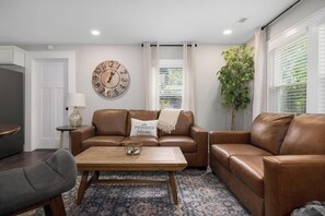 A living room with a brown leather sofa, a wooden coffee table, a round wall clock, a potted plant, and a window with blinds.
