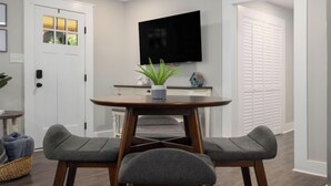A dining area with a round table, two grey upholstered chairs, a potted plant, a flat-screen TV, and a white door. - Walk to Downtown Jboro · Remodeled 3BR · Sleeps 6 (Jonesborough)