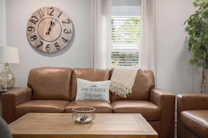 Un salon avec un canapé en cuir marron, une table basse en bois, une horloge murale et une fenêtre avec des stores.