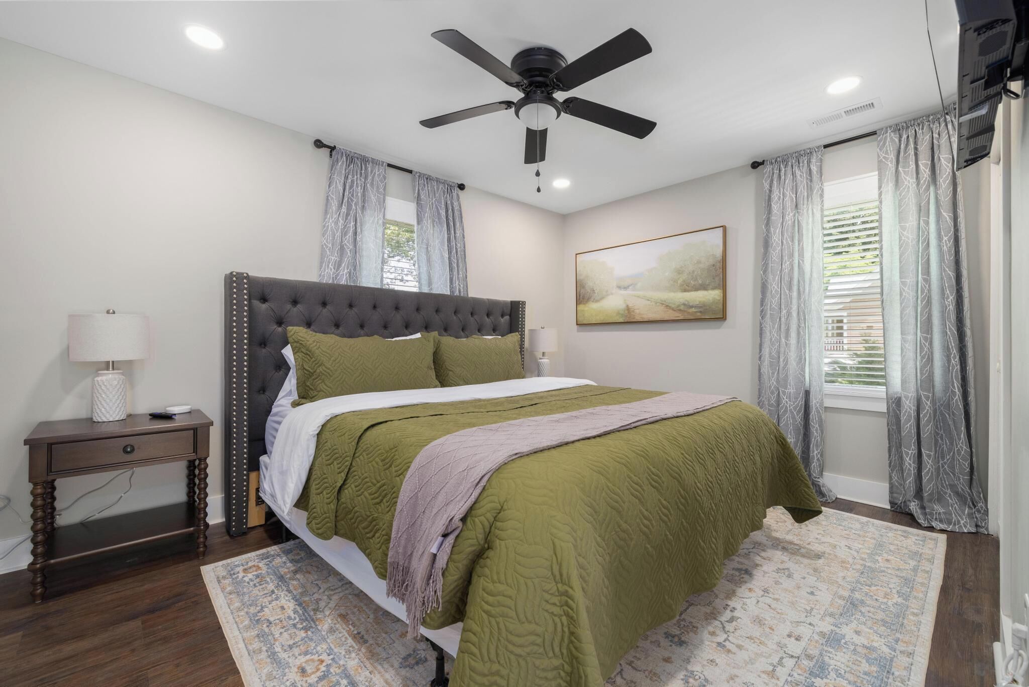A bedroom with a tufted headboard, a bed with green bedding, a bedside table with a lamp, and a ceiling fan.