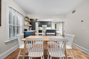 A dining area with a wooden table and white chairs, a kitchen in the background, and a window with blinds.