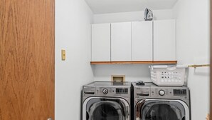 Une buanderie équipée de deux machines à laver et deux sèche-linge à chargement frontal, de placards en bois au-dessus et d’une petite étagère où sont rangés des paniers à linge.