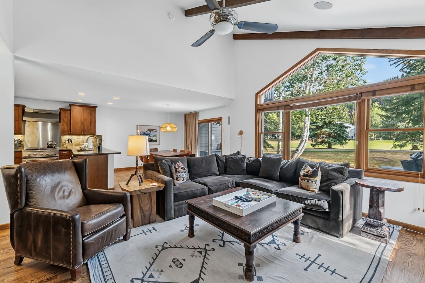 A spacious living room with a large sectional sofa, a leather armchair, and a coffee table. There is a dining area in the background with a kitchen visible.