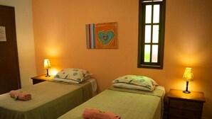 A room with two beds, each with a pillow and a rolled towel. There is a window with blinds, a wall-mounted artwork, and a nightstand with a lamp. - MOANA  (Buzios)